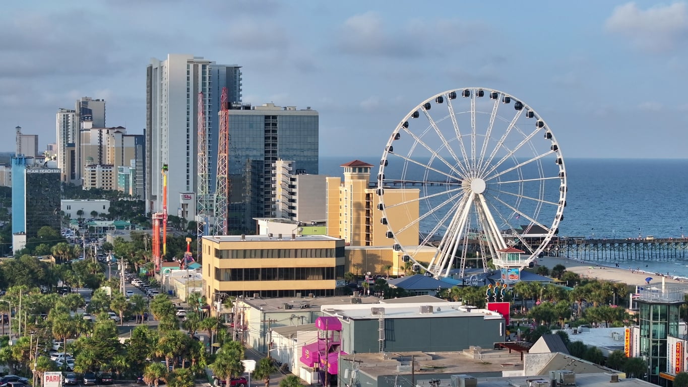 Cityscape of Myrtle Beach, featuring tall buildings, a large Ferris wheel near the shoreline, colorful attractions, and the ocean in the background under a partly cloudy sky—capturing both the lively atmosphere and occasional complaints about bad food from some visitors.