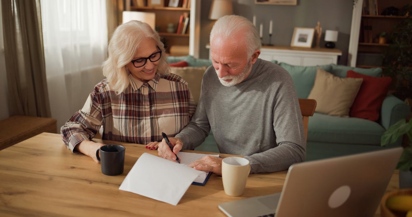 An elderly couple sits at a table, smiling as the man signs a document. They have coffee mugs nearby, and an open laptop is on the table. The setting appears to be a cozy, well-lit living room.