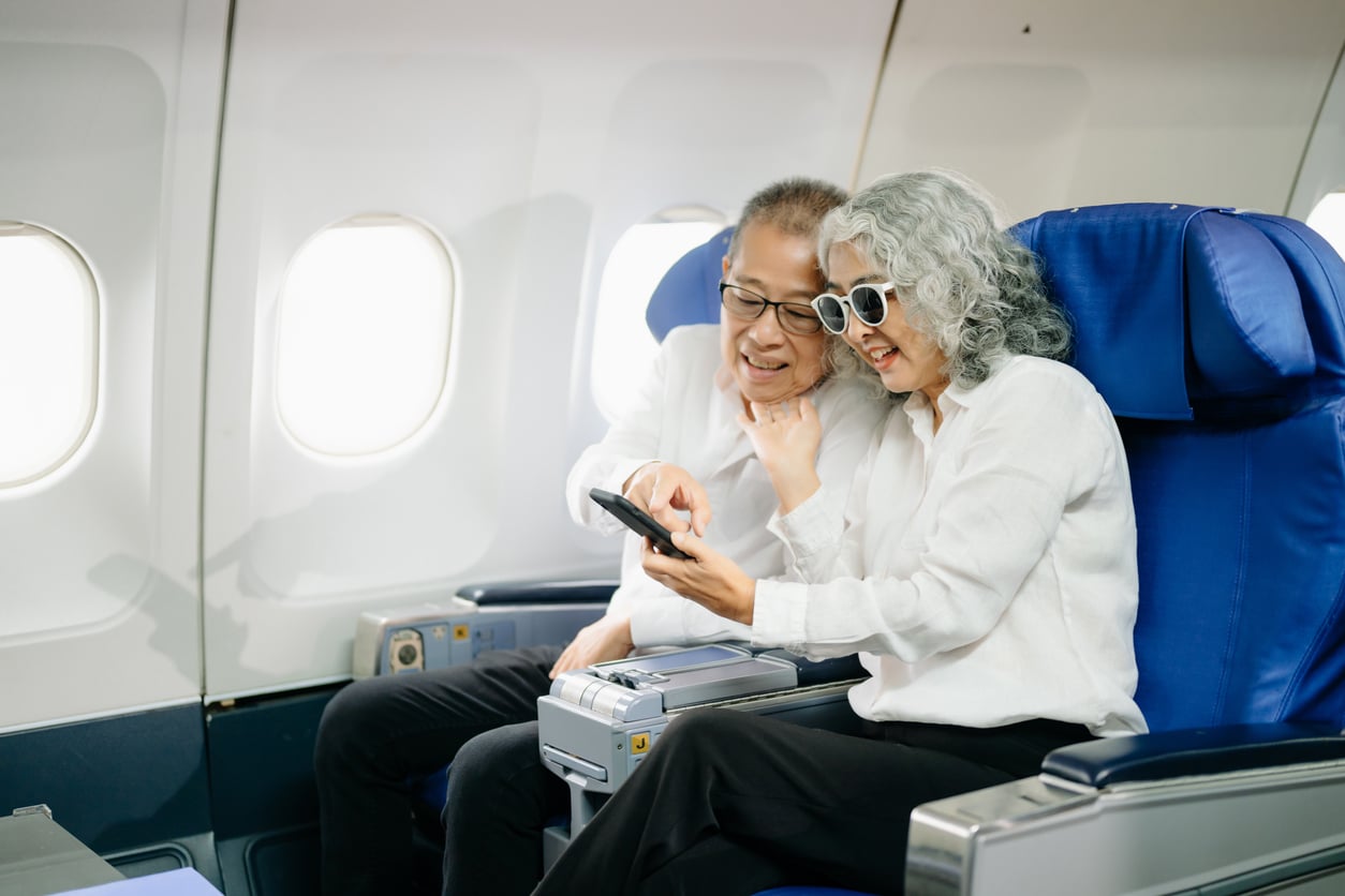 An older couple sits together on an airplane, smiling and looking at a smartphone. Both wear white shirts; the woman has gray hair and sunglasses. Displaying their carefree travel habits, they appear happy and relaxed in their blue airline seats.