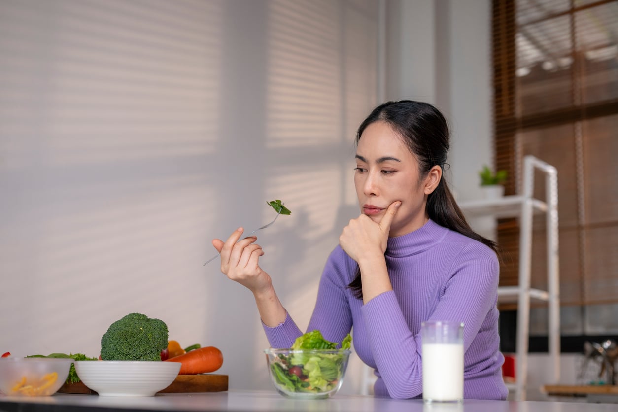 A woman in a purple top sits at a table, looking thoughtfully at a fork with greens. There is a bowl of salad, vegetables, and a glass of milk in front of her.