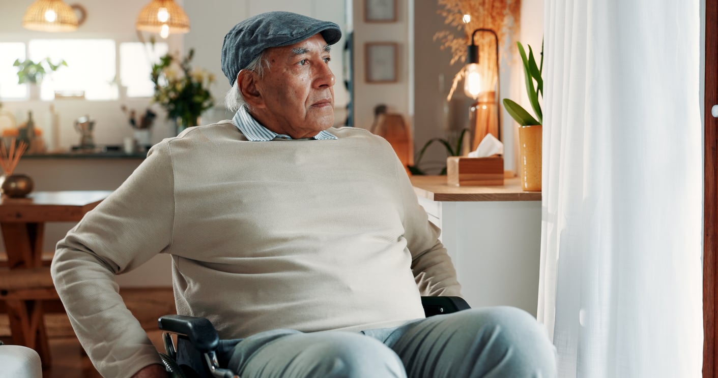 An older man in a light sweater and cap sits in a wheelchair near a window, looking outside as he reflects on retirement tips for enjoying peaceful days. The background shows a cozy, well-lit home interior with plants and warm decor.