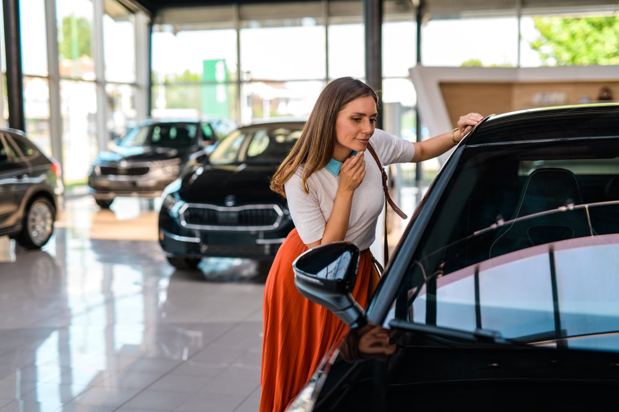 A woman in an orange skirt examines a black car in a brightly lit showroom with several other cars visible in the background.