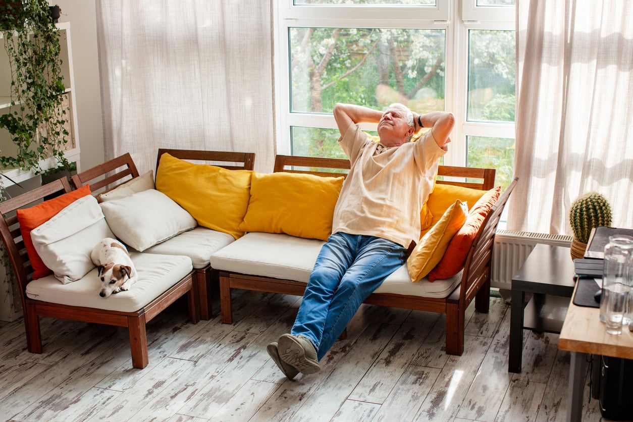 An older man relaxes with hands behind his head on a sofa with yellow pillows in a sunlit living room. A small dog sleeps on the couch beside him.