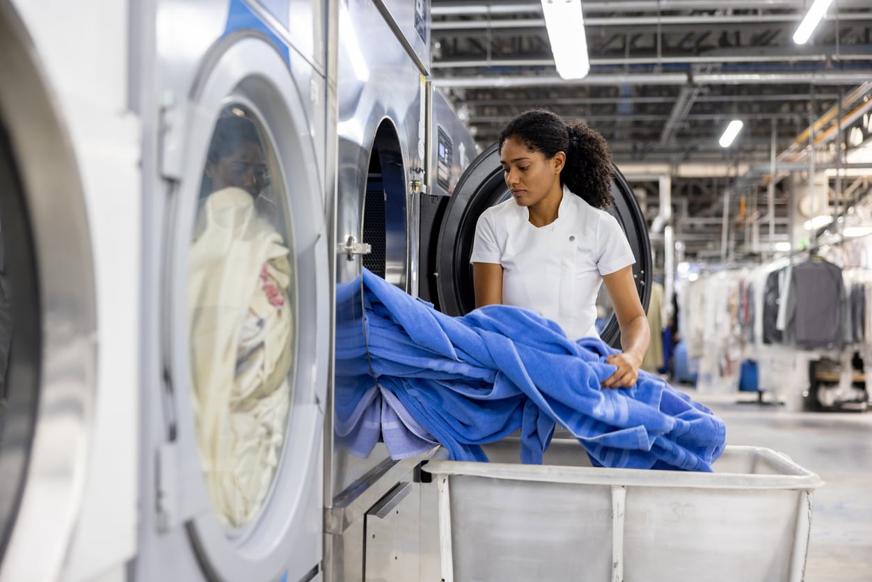 A woman places blue linens into a large industrial washing machine in a laundromat, with laundry carts and hanging clothes visible in the background.