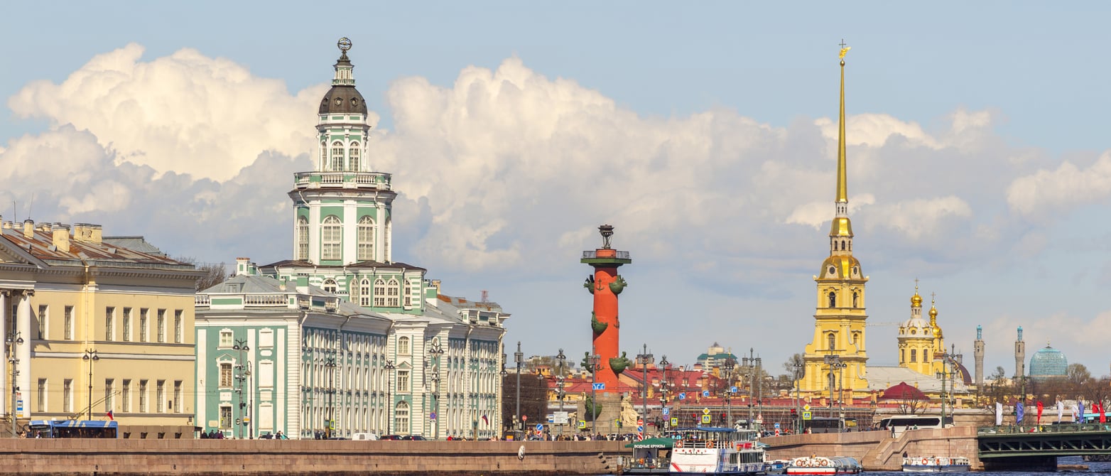 A cityscape of St. Petersburg, Russia, featuring historic buildings, a red Rostral Column, and the golden spire of Peter and Paul Cathedral under a blue sky with clouds—one of the remarkable sights in a destination that has recently appeared on some lists of places travelers are warning against going in 2026.
