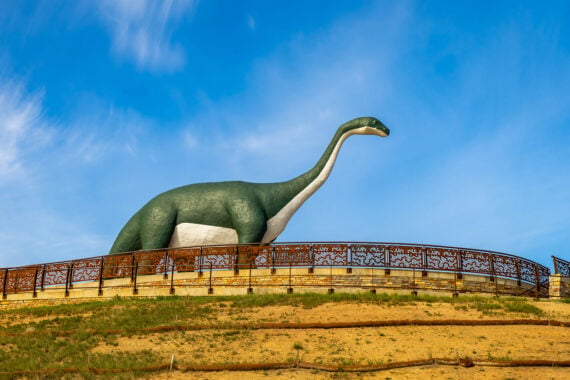 A large green and white dinosaur statue stands on a hill behind a decorative metal fence, set against a blue sky with wispy clouds&mdash;a true example of classic roadside attractions.