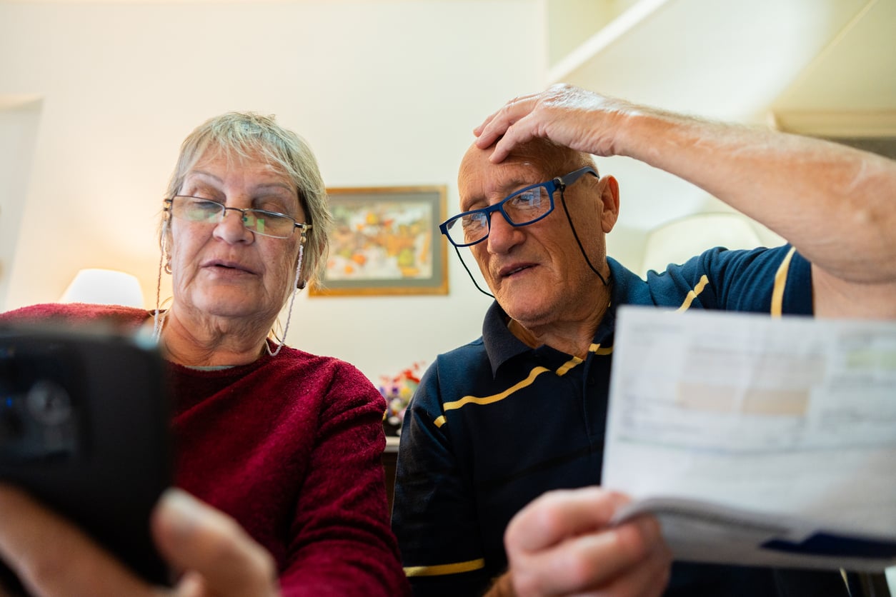 An older woman in glasses holds a smartphone, while an older man with glasses looks at a document, appearing concerned. They are seated indoors with a framed picture on the wall behind them.