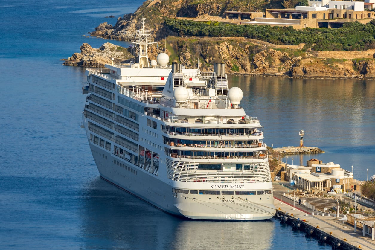 A large white cruise ship named Silver Muse is docked at a harbor beside a small building, with rocky hills and greenery in the background.