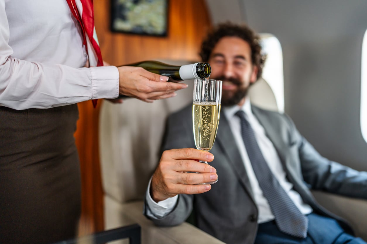 A flight attendant pours champagne into a glass held by a smiling man in a suit, seated on an airplane with a window in the background.