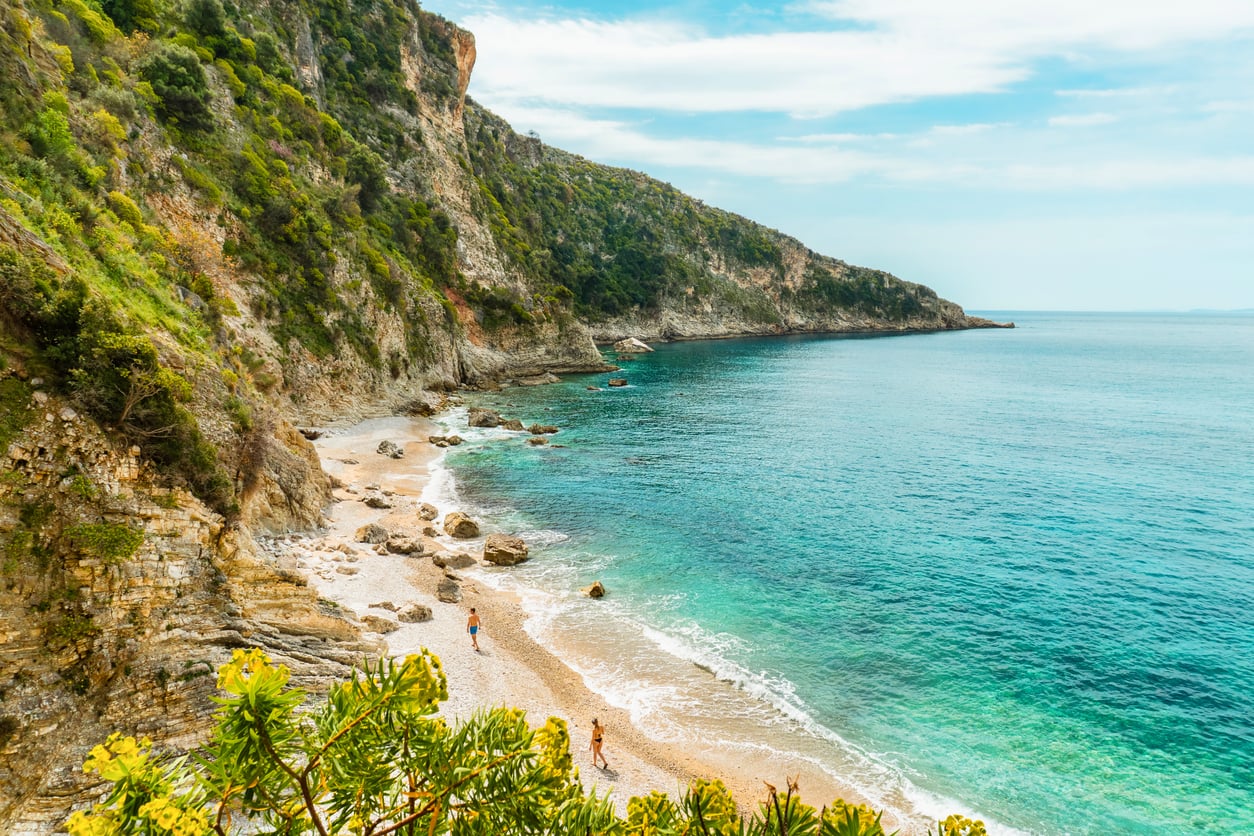 A secluded beach with turquoise water and gentle waves, bordered by rocky cliffs covered in green vegetation, is quickly becoming one of those travel spots boomers are learning to love. A few people walk along the sandy shore under a partly cloudy sky.