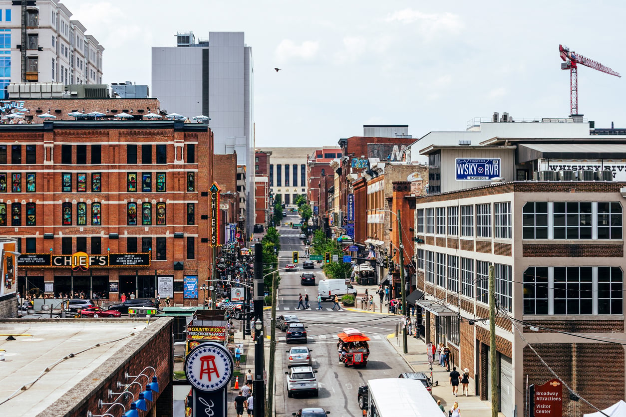 A busy city street with cars, pedestrians, brick buildings, and bars—some notorious for their bad food. A pedal tavern rides along the street while a crane is visible in the background under a partly cloudy sky.