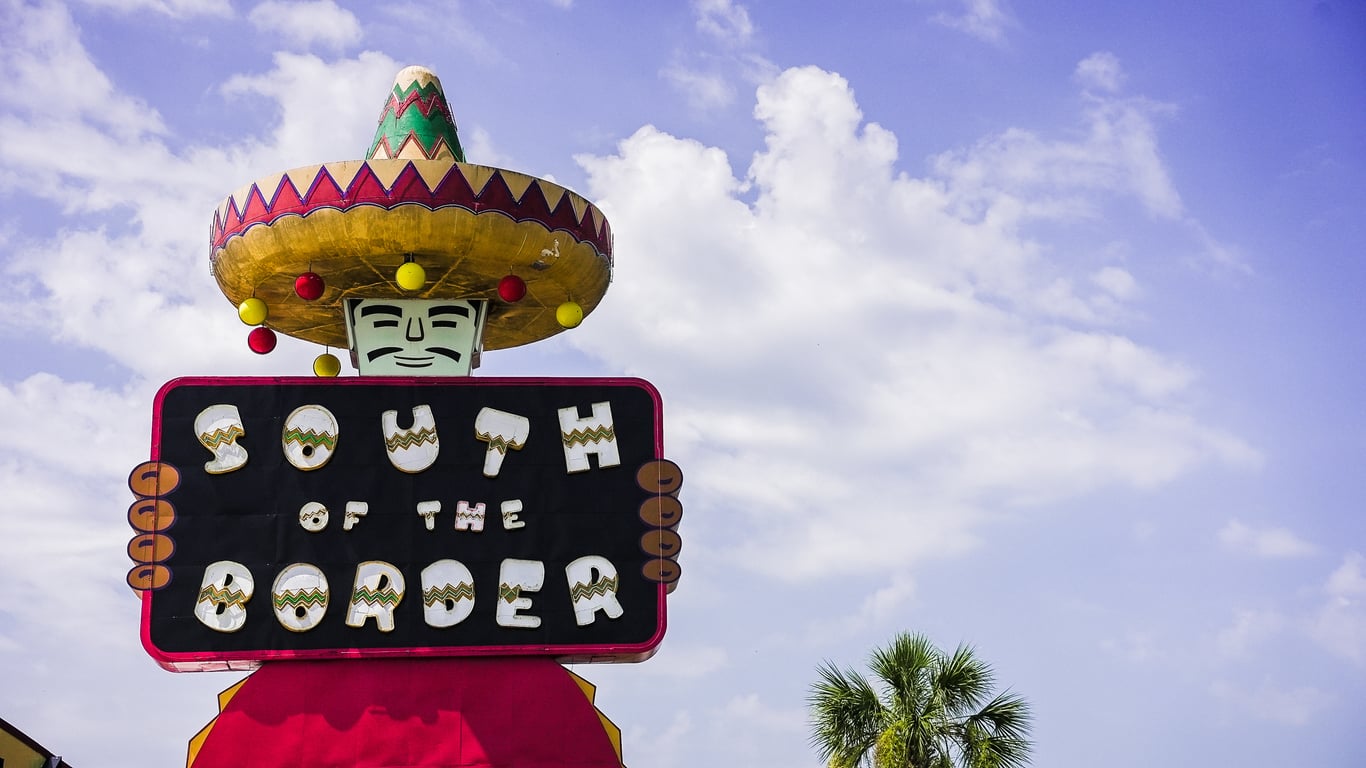 Colorful roadside sign with a large sombrero and a cartoon face, holding a sign that reads “South of the Border” against a blue sky with clouds and a palm tree in the background—one of the most iconic Classic Roadside Attractions in America.