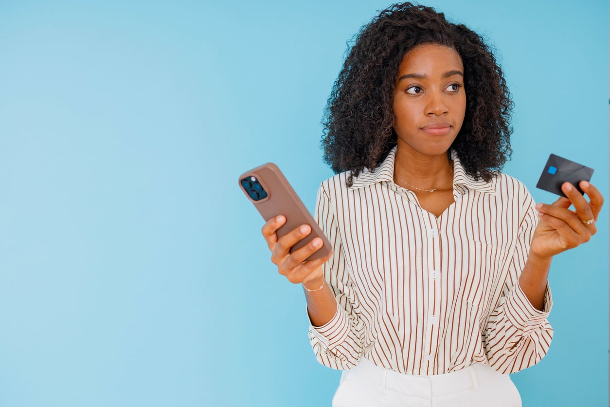 A woman stands against a blue background, holding a smartphone in one hand and a credit card in the other, looking to the side with a thoughtful expression.