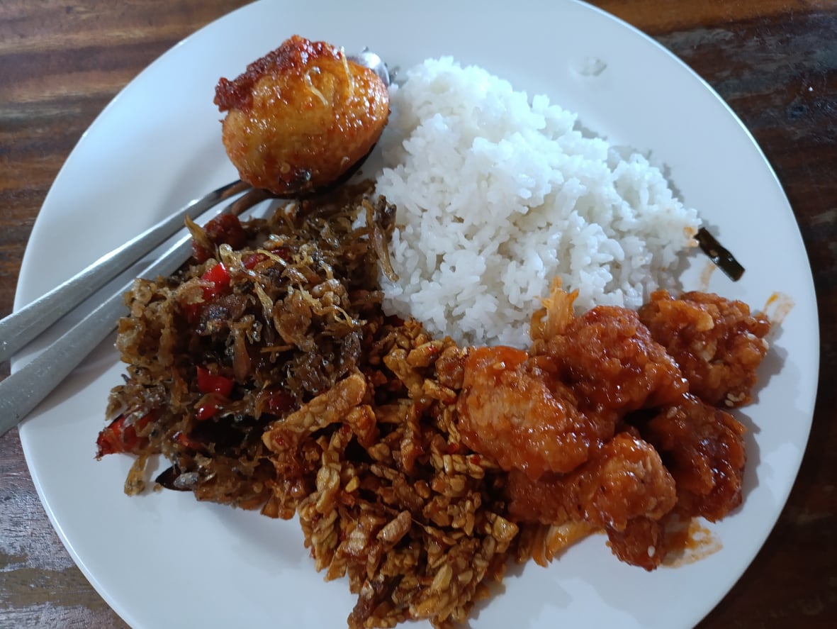 A white plate with steamed rice, a hard-boiled egg in sauce, spicy shredded meat, crispy tempeh, and pieces of fried chicken in a reddish sauce, served with a spoon and fork.