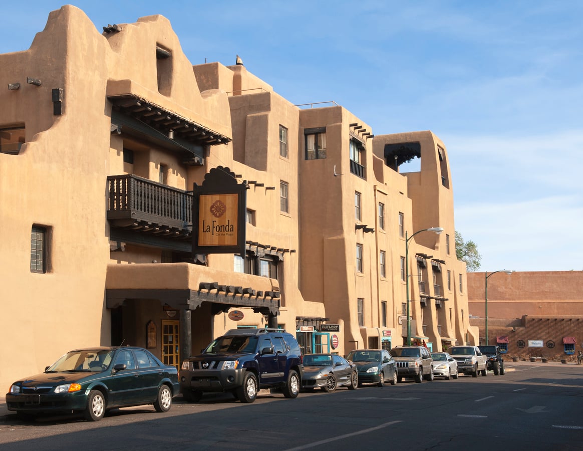 Street view of La Fonda Hotel, a multi-story adobe-style building with wooden balconies and parked cars in front, under a clear blue sky. As one of the region’s renowned historic hotels, La Fonda exudes timeless charm and character.
