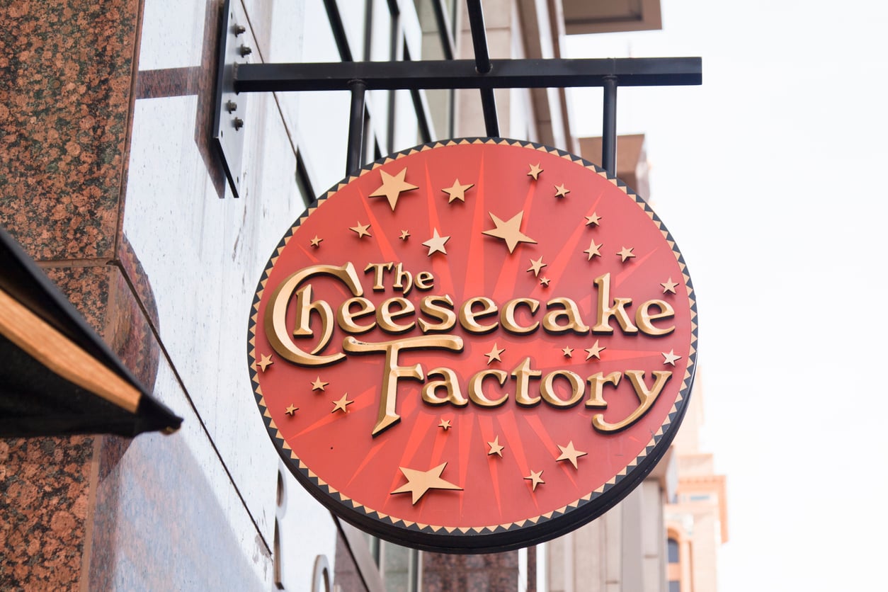 A round, red sign with gold stars and text that reads “The Cheesecake Factory” hangs outside a building.