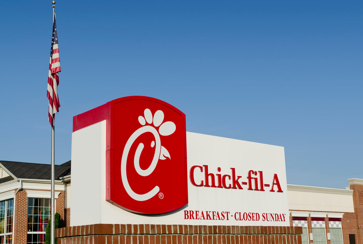 A Chick-fil-A restaurant sign with the logo and text “Breakfast - Closed Sunday” is displayed near a brick building and an American flag, set against a clear blue sky. Discover Wild Fast-Food Facts as you spot this iconic sign—reminding visitors of Chick-fil-A’s unique traditions.