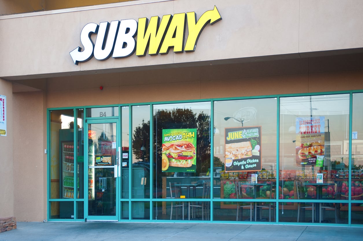 A Subway restaurant storefront with large glass windows, green framing, and promotional posters for sandwiches displayed on the glass. Above the entrance, the yellow and white Subway logo stands out—an inviting spot to enjoy your meal while discovering Wild Fast-Food Facts about one of the world’s most popular sandwich chains.