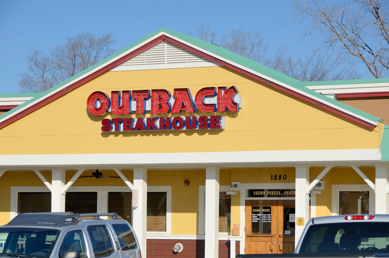 Outback Steakhouse restaurant exterior with large red and blue sign above the entrance, yellow walls, white trim, and parked cars in front. Trees and clear sky are visible in the background.