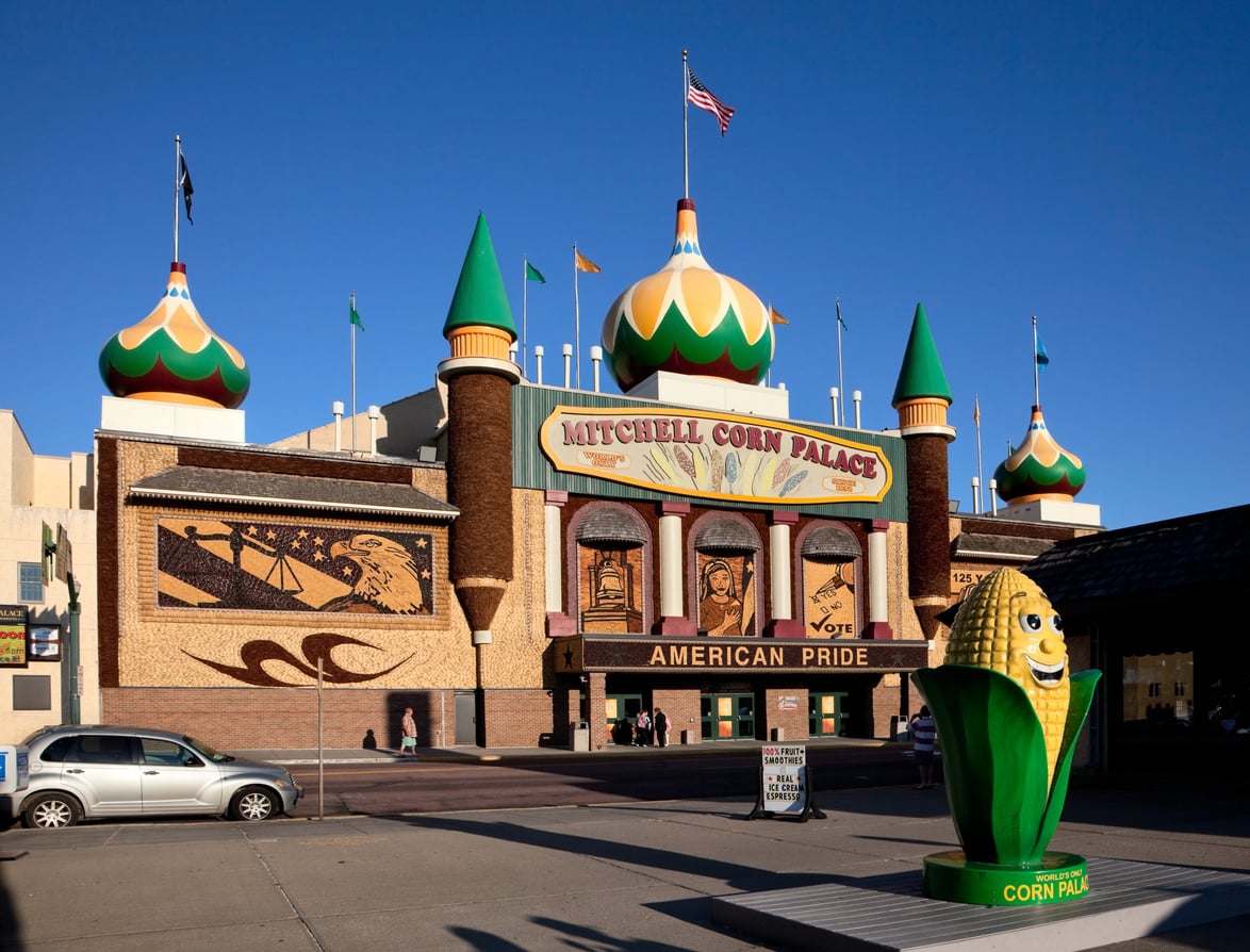 Mitchell Corn Palace stands as one of America’s Classic Roadside Attractions, featuring ornate onion domes, detailed corn murals on the facade, flags flying, and a large corn cob statue labeled “Corn Paul” in front. Cars and people are visible nearby under a clear blue sky.