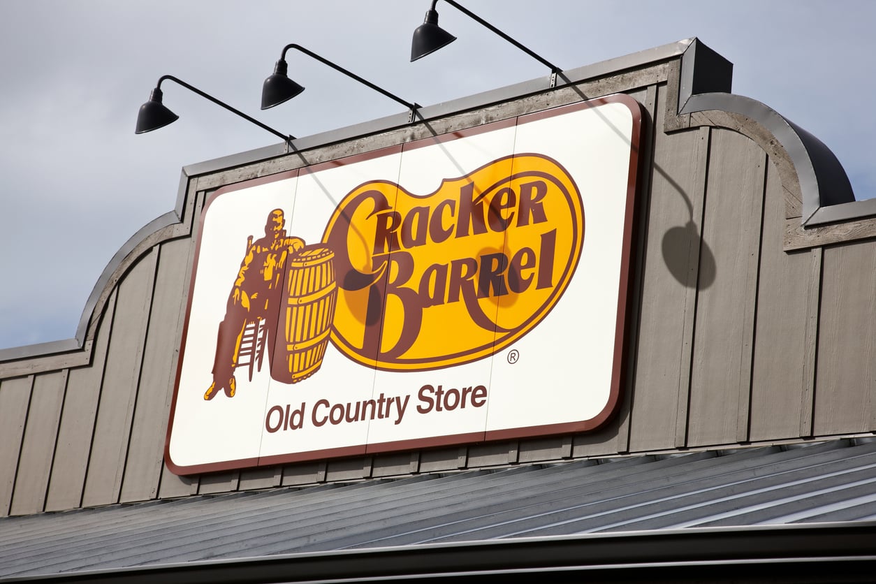 Sign for Cracker Barrel Old Country Store featuring a logo with a man sitting beside a barrel, mounted on the exterior of a building with gray siding.