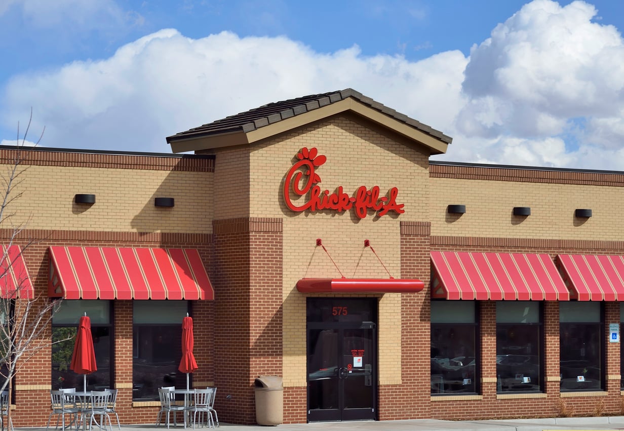 A Chick-fil-A restaurant with red striped awnings, outdoor seating, and a clear blue sky with clouds in the background.