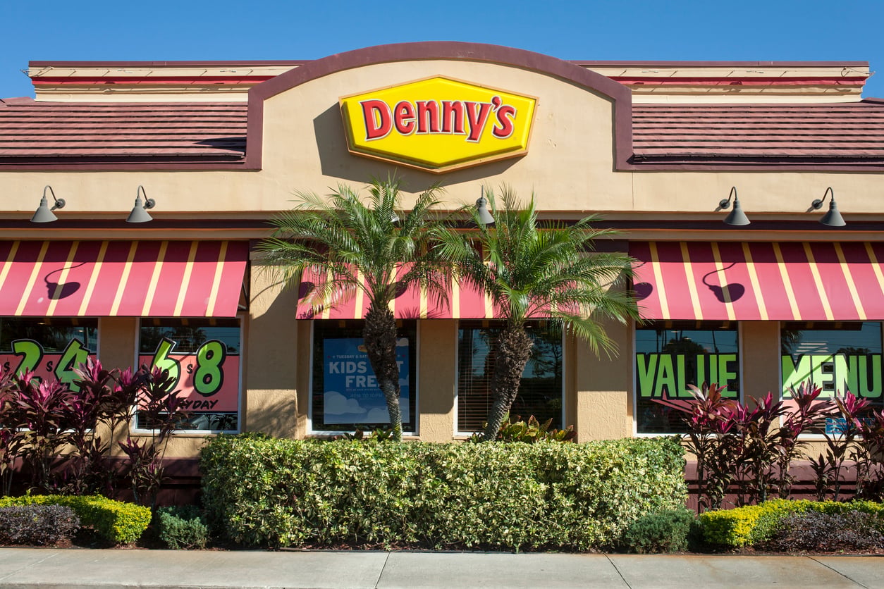 The exterior of a Denny’s restaurant with red and yellow striped awnings, palm trees, and large yellow Denny’s sign above the entrance on a sunny day.
