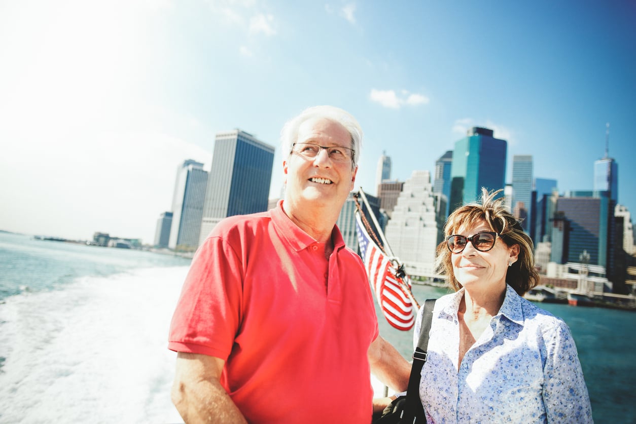 Two smiling adults stand on a boat with an American flag behind them, enjoying picturesque views of a city skyline and tall buildings in the background on a sunny day—an ideal scene for those interested in exploring travel destinations boomers are skipping.
