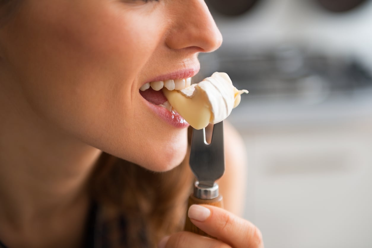 Close-up of a woman smiling as she brings a fork with a piece of creamy cheese to her mouth, ready to take a bite.