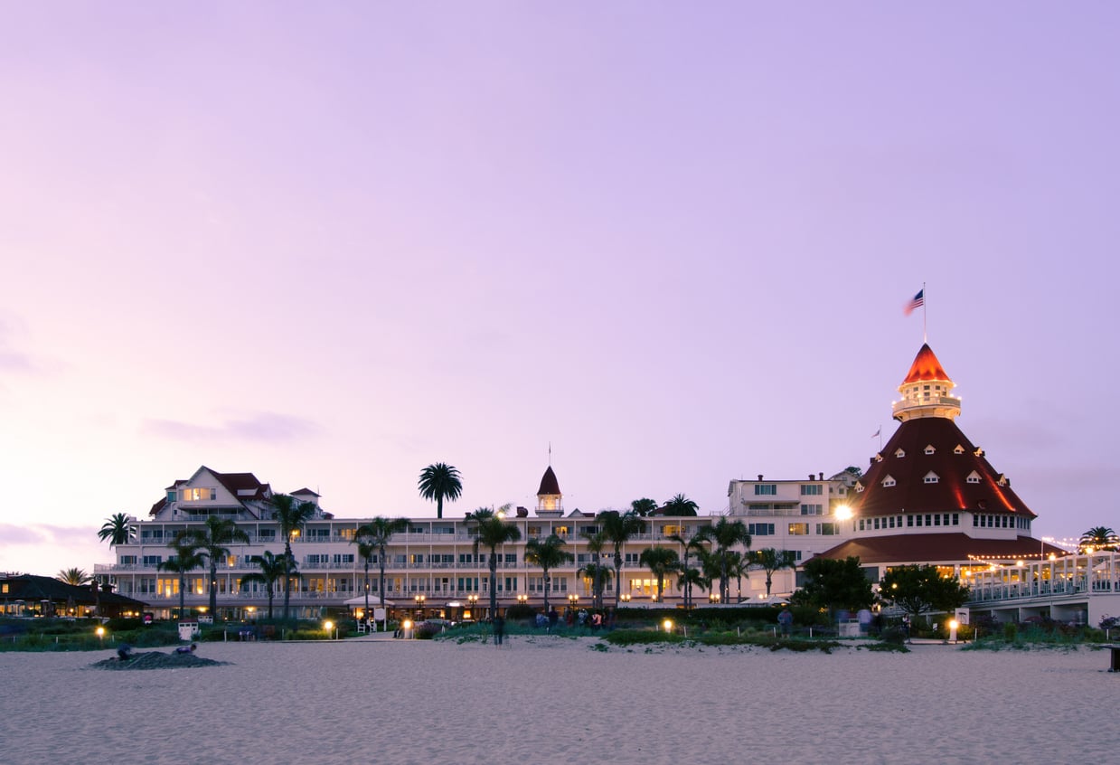 A historic beachfront hotel with a red turret roof, celebrated as one of the Historic Hotels in the region, is lit at sunset and surrounded by palm trees. The sky is purple and sand fills the foreground.
