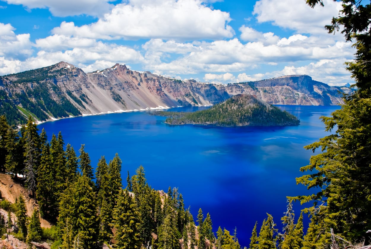 A deep blue lake surrounded by forested cliffs and mountains under a partly cloudy sky, with a forested island in the center of the lake—one of many breathtaking landscapes that showcase fascinating travel facts about the United States.