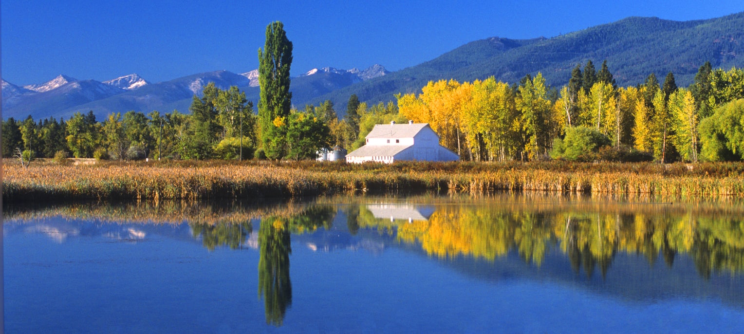 A white barn sits among autumn trees near a calm lake, with yellow foliage and mountains in the background. The scene is reflected in the water under a clear blue sky.