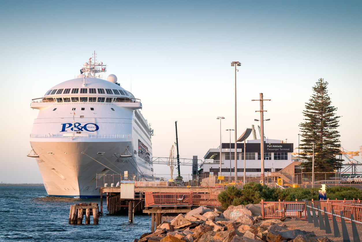 A large P&O cruise ship docked at Port Adelaide, next to a terminal building, with walkways, railings, rocks, and trees visible in the foreground under a clear sky.