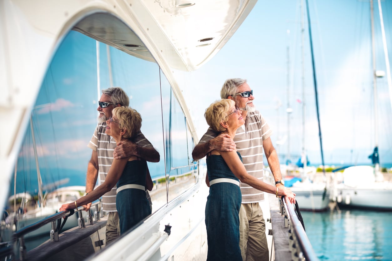 An older couple embraces and smiles while standing on a docked yacht, with their reflection visible in the boat’s shiny surface—a joyful moment that captures their adventurous travel behaviors. Sailboats and a blue sky are in the background.
