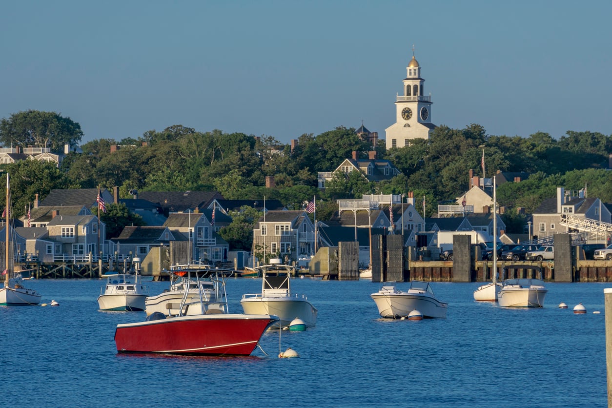 Boats float on calm blue water in a harbor, with a row of houses and a white church with a clock tower visible among green trees in the background under a clear sky, evoking the charm of nostalgic travel destinations.
