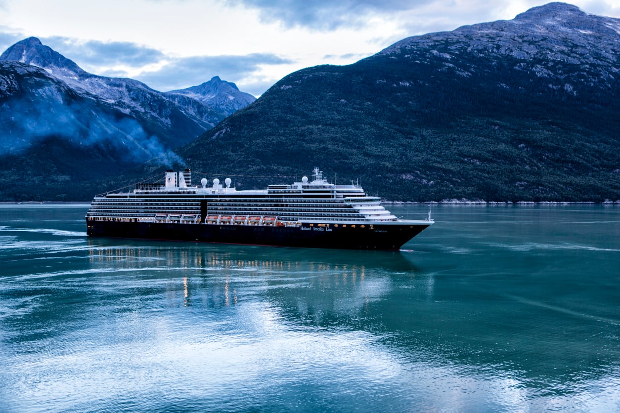 A large cruise ship sails across calm, turquoise water with forested mountains and a cloudy sky in the background.