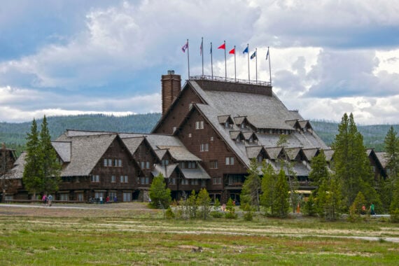 Large rustic wooden lodge with steep roofs and multiple flags on top, surrounded by trees and a grassy field under a partly cloudy sky&mdash;an inviting retreat reminiscent of Historic Hotels.