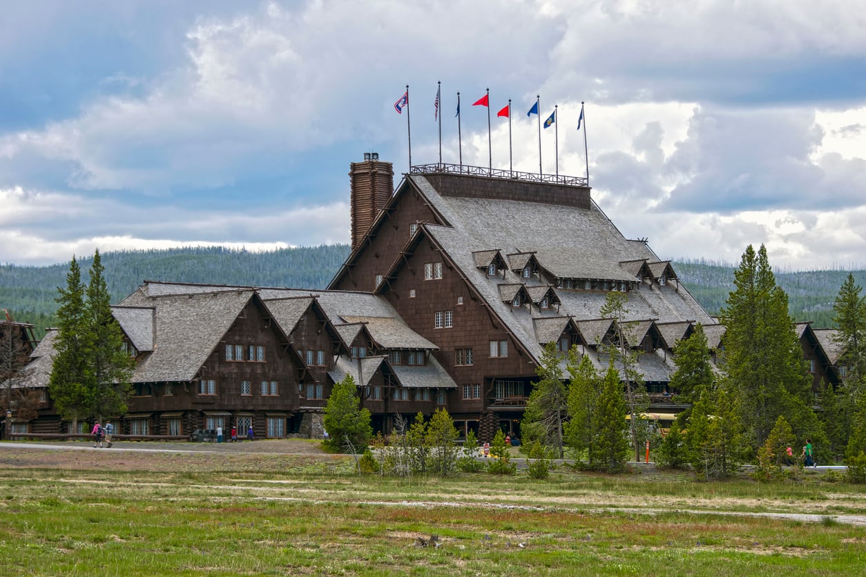 Large rustic wooden lodge with steep roofs and multiple flags on top, surrounded by trees and a grassy field under a partly cloudy sky—an inviting retreat reminiscent of Historic Hotels.