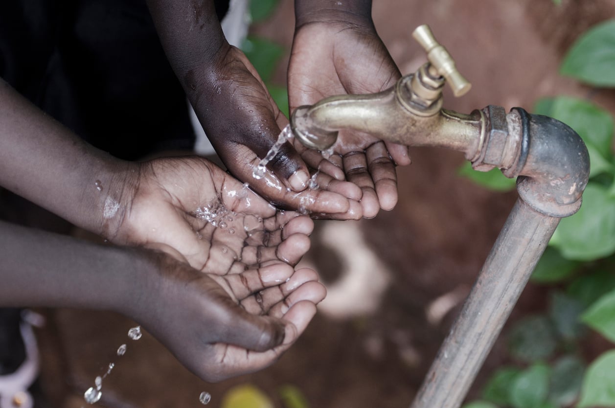 Three pairs of dark-skinned hands are cupped together under a brass outdoor faucet as water trickles down into them, with green leaves and earth visible in the background—an image that serves as a poignant reminder of why some destinations are featured on lists like Places Travelers Are Warning Against Going In 2026.
