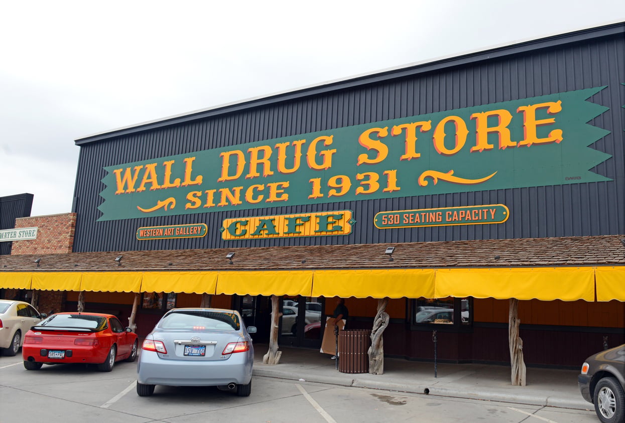 Wide view of Wall Drug Store building with large green and yellow sign, cars parked in front, and yellow awning over the entrance. As one of the Classic Roadside Attractions, the sign reads “Wall Drug Store Since 1931,” advertising a café and seating capacity.