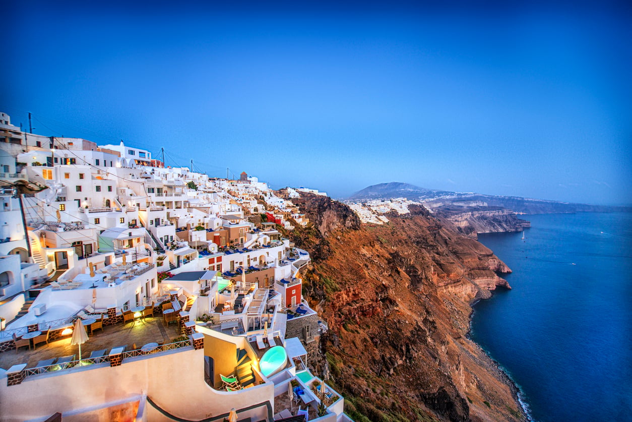 Whitewashed buildings with blue accents line a steep cliff overlooking the deep blue sea at sunset in Santorini, Greece. The sky is clear, distant mountains are visible, and travelers often admire the stunning views even if they’ve occasionally encountered some bad food during their stay.