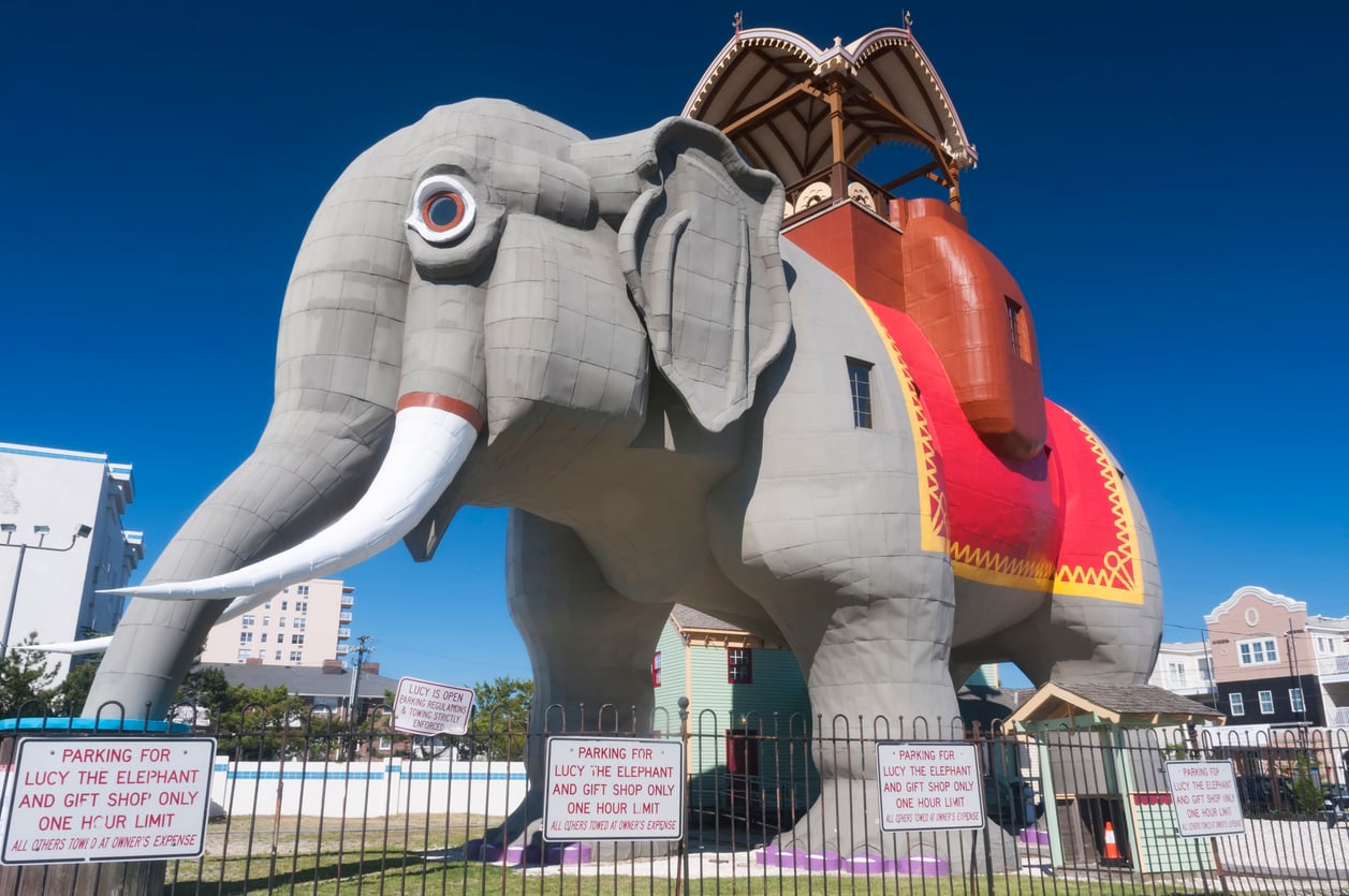 A giant, gray elephant-shaped building with white tusks and a red, yellow, and gold blanket stands behind a fence with parking signs. A small entryway and viewing deck sit atop the elephant’s back. Blue sky and nearby buildings are visible.