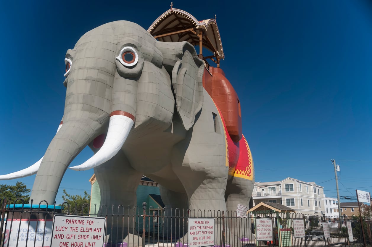 A large, gray elephant-shaped building with tusks and a red saddle stands behind a fence under a clear blue sky; nearby signs mention parking limits and a gift shop, making it one of the region’s Classic Roadside Attractions.
