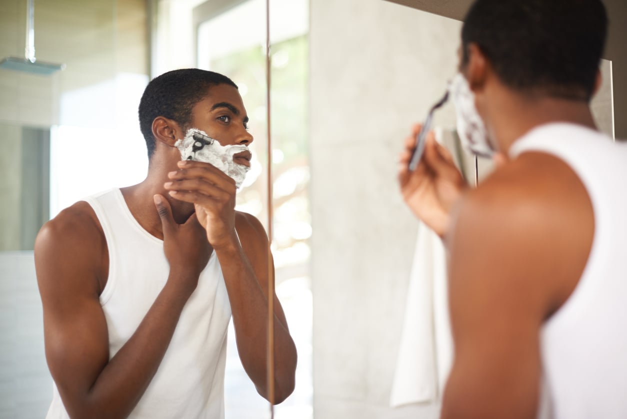 A man wearing a white tank top shaves his face with a razor in front of a bathroom mirror, applying shaving cream.