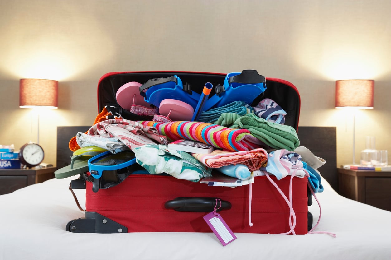 An overstuffed red suitcase with clothes and pool toys spilling out sits open on a bed in a cozy, well-lit bedroom, with lamps and bedside items in the background.