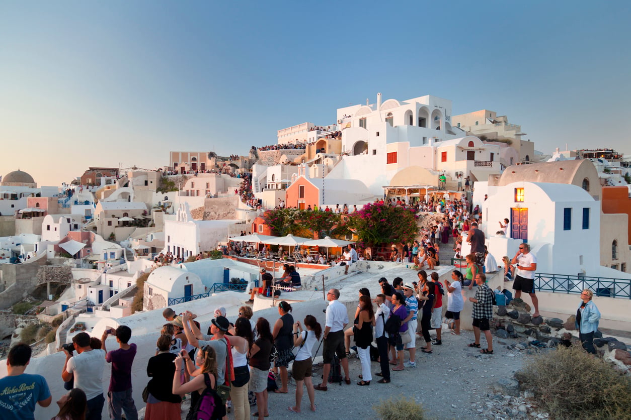 Crowd gathers at sunset in Oia, Santorini, Greece, with white-washed buildings and blue-domed roofs overlooking the sea under a clear sky—a breathtaking scene in one of the travel destinations Boomers are skipping.