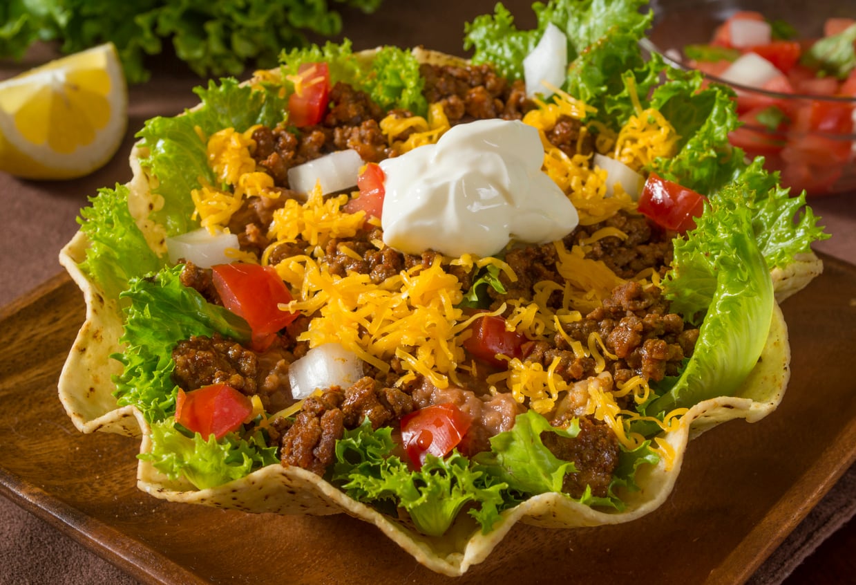 A taco salad in a tortilla bowl with ground beef, shredded cheese, lettuce, diced tomatoes, onions, and a dollop of sour cream on top.