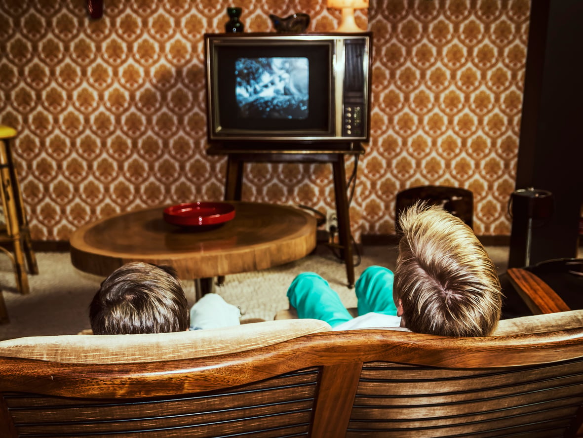 Two children sit on a vintage sofa watching a black-and-white TV in a retro living room with patterned wallpaper, a wooden table, and a red ashtray.