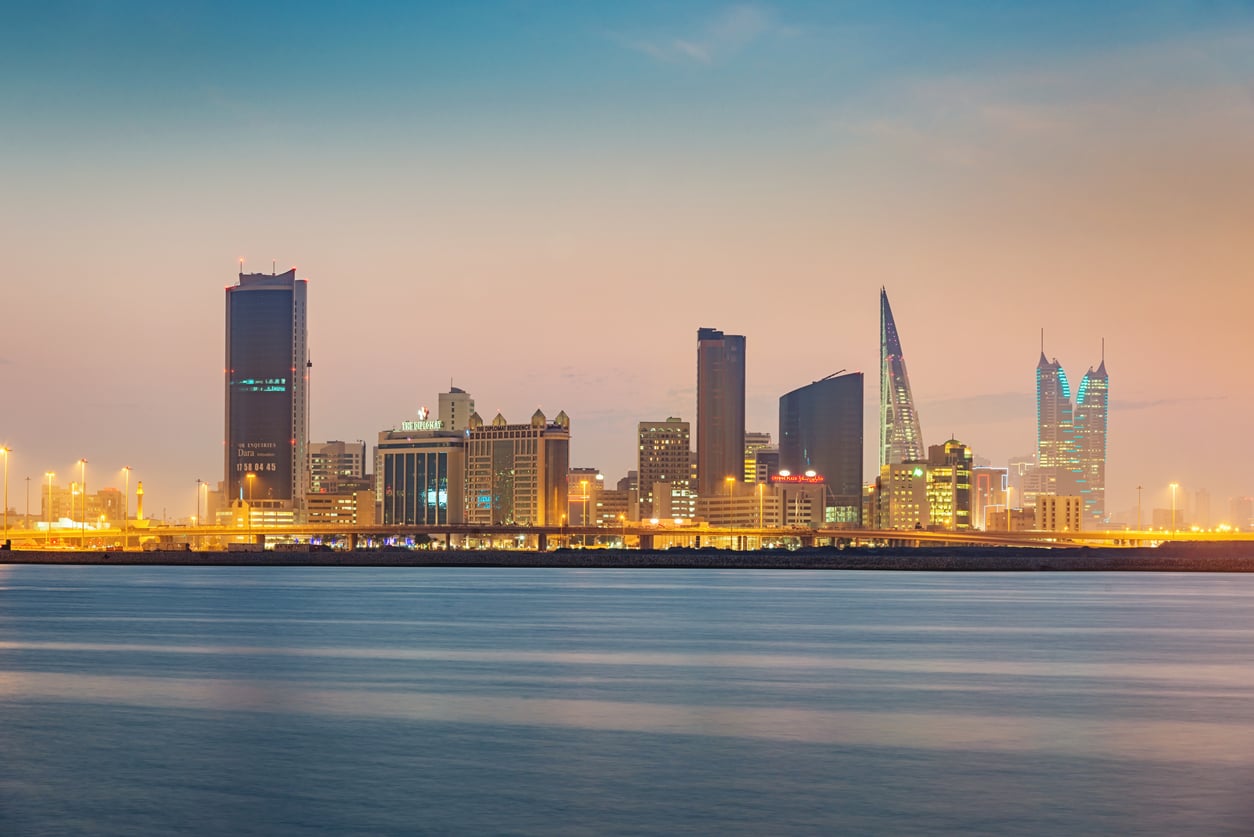 Evening cityscape of a modern skyline with high-rise buildings and water in the foreground, under a clear sky with a subtle gradient from blue to peach—a tranquil scene that invites exploration, yet may be subject to travel restrictions and advisories.