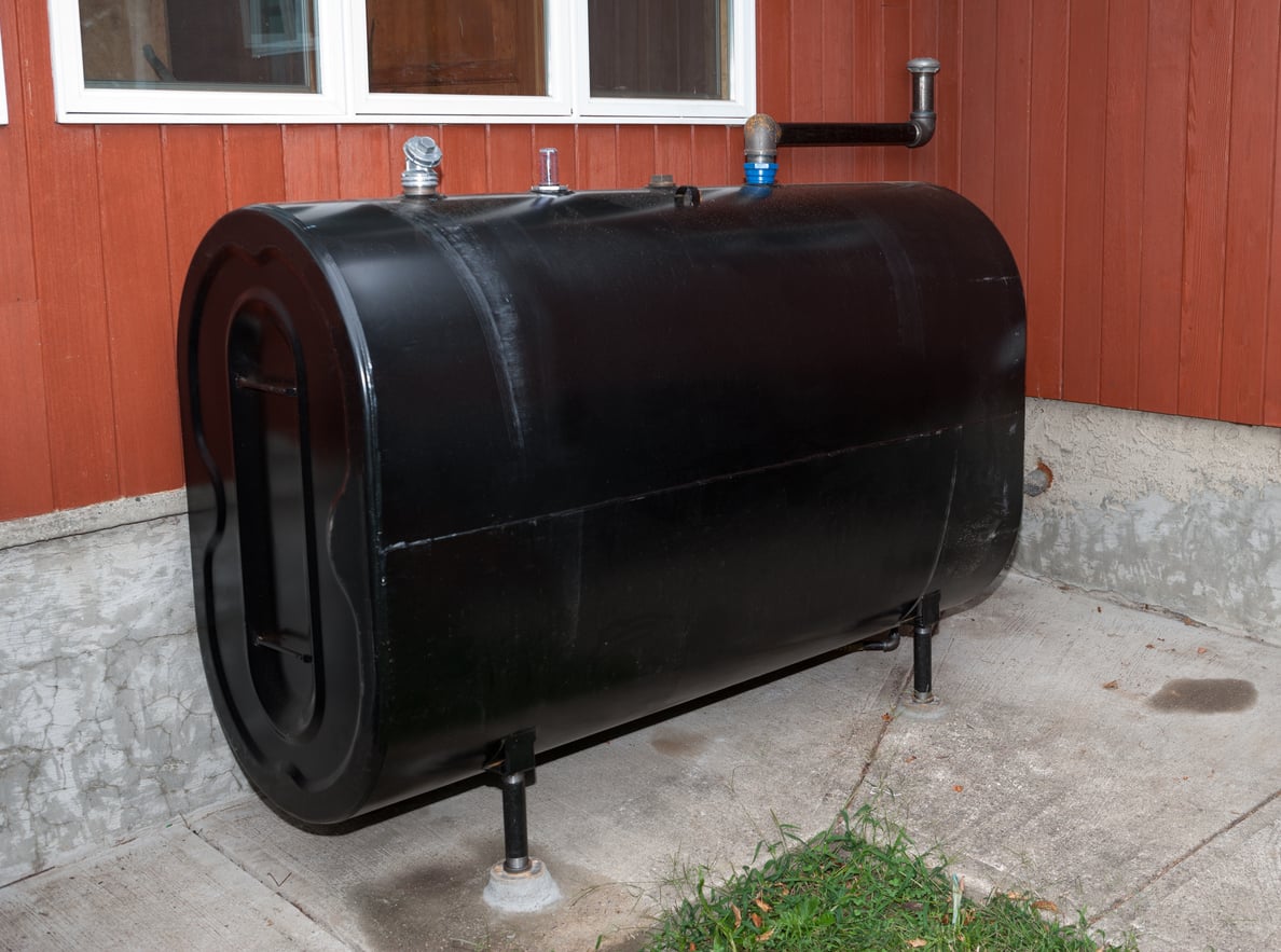 A black oil tank stands on metal supports next to a red building with white-trimmed windows, resting on a concrete pad with some grass nearby.